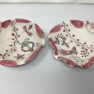 May include: Two small, white ceramic dishes with pink and black painted designs. One dish has a lyre design and the other has a floral design.