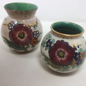May include: Two ceramic vases with a round shape and a wide opening. The vases are decorated with a large burgundy flower, green leaves, and smaller blue, orange, and yellow flowers. The interior of the vases is green.