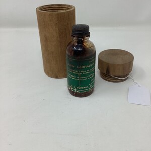 May include: A small brown glass bottle with a black cap and a green label, next to a wooden container and a wooden lid. The label reads "New Labrador". A blank white tag is attached to the lid.