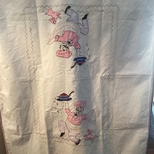 May include: White tablecloth with embroidered figures of chefs in pink and white checkered outfits. One chef is upside down, and the other is holding a plate of food. A pink pig is also embroidered on the tablecloth.