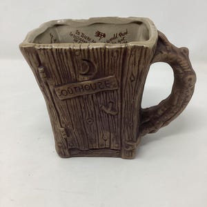 May include: A brown ceramic mug with a tree trunk design, featuring a branch-shaped handle. The mug has the word "OUTHOUSE" on the front and text inside the rim. The mug is square-shaped and has a rustic appearance.