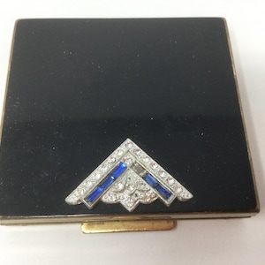 May include: A vintage black compact with a decorative silver and jewel-toned accent. The accent features clear and blue rhinestones in a geometric design. The compact has a gold-toned metal frame and clasp.