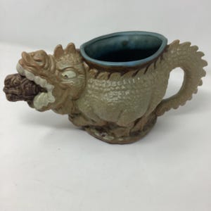 May include: A ceramic dragon-shaped pitcher with a handle and a spout. The dragon is primarily tan and green, with a blue interior and a brown object in its mouth. The pitcher has a textured, scaly appearance and is set against a white background.