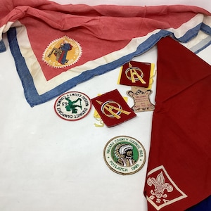 May include: A collection of vintage Boy Scout patches and neckerchiefs. The patches feature various designs, including a scout in a red uniform, a compass, and a Native American headdress. The neckerchiefs are red and blue with white trim.