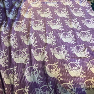 May include: A purple fabric with a repeating pattern of white floral baskets and flowers. The fabric has a scalloped edge and a dotted background. The design is a vintage style, suitable for home decor or crafting projects.