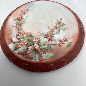 May include: Round, antique ceramic plate with a red rim and a floral design. The plate features a hand-painted design of red berries and green leaves on a cream and peach-colored background.