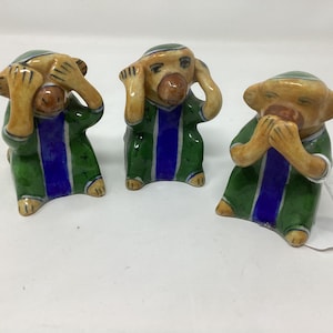 May include: Three ceramic monkey figurines, each with a different gesture: one covering its eyes, one covering its ears, and one covering its mouth. The monkeys are painted in shades of green, blue, and yellow.