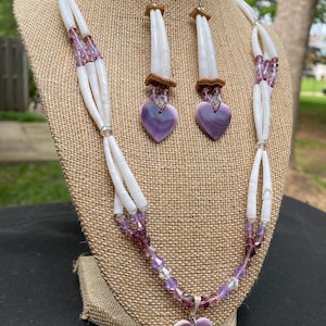 May include: A white beaded necklace and matching earrings set. The necklace features strands of white beads interspersed with purple and clear crystal beads, and a purple heart pendant. The earrings have a similar design, with purple heart-shaped pendants.