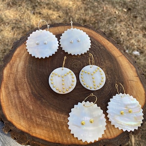 Op de afbeelding: Een collectie handgemaakte oorbellen in diverse ontwerpen. De oorbellen zijn gemaakt van witte, iriserende schelpen met zilveren of goudkleurige accenten. Sommige oorbellen hebben een geschulpte rand, andere zijn cirkelvormig. De afbeelding bevat de tekst "@beadinbycourt".