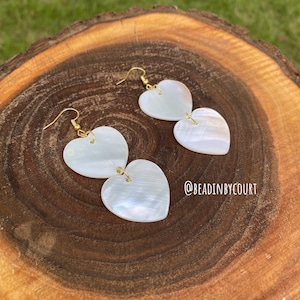 Peut inclure: Boucles d'oreilles en forme de cœur avec une finition nacrée, reliées en cascade. Les boucles d'oreilles sont suspendues à des crochets dorés. Les boucles d'oreilles sont présentées sur une surface en bois avec un grain naturel.