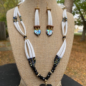 Könnte beinhalten: Ein weißes Muschelkette und passendes Ohrring-Set. Die Halskette hat einen herzförmigen Anhänger mit schillernder Abalone-Muschel. Die Ohrringe haben herzförmige Abalone-Muschel-Anhänger und braune Lederakzente. Das Set wird auf einer Jutefläche präsentiert.