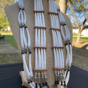 May include: A white shell necklace and matching earrings set. The necklace features multiple strands of white shells, accented with black and clear beads. The earrings have a similar design, with brown accents. The set is displayed on a mannequin.