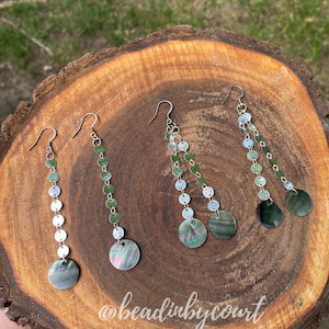 May include: Four pairs of dangle earrings with silver hooks and chains. The earrings feature small, round, green and silver beads, and a larger, iridescent, circular charm at the bottom. The earrings are displayed on a wooden surface. The text "@beadinbycourt" is visible.