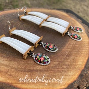 Könnte beinhalten: Handgefertigte Ohrringe mit weißen, geschichteten Muschelsegmenten und ovalen Anhängern mit floralen Designs. Die Anhänger haben einen schwarzen Hintergrund mit roten, rosa und grünen Blumen und einen silberfarbenen Rand.