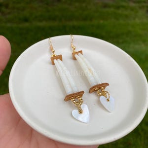 May include: A pair of white shell earrings with gold accents and heart-shaped charms. The earrings are hanging from a small plate.