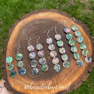May include: A collection of abalone shell earrings with silver hooks, presented on a wooden board. Each earring features multiple iridescent, circular shell pieces, displaying colours like green, blue, and purple. The earrings are a decorative accessory.