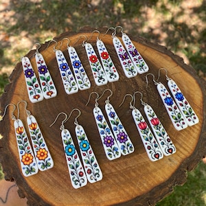Può includere: Una collezione di orecchini pendenti rettangolari bianchi con motivi floreali colorati, steli e foglie verdi. Ogni paio presenta distinti disegni di fiori nelle tonalità del rosso, blu, viola, arancione e giallo, con ganci color argento.
