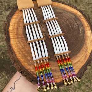 Pode incluir: Um par de brincos de osso branco com contas de cristal arco-íris e sinos dourados. Os brincos são presos a tiras de couro marrom. @beadinbycourt