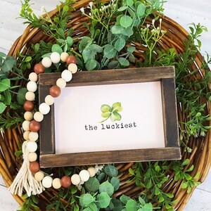May include: A wooden sign with the text "the luckiest" and a watercolor illustration of a four-leaf clover. The sign is surrounded by a wreath of green leaves and a strand of wooden beads with a tassel.