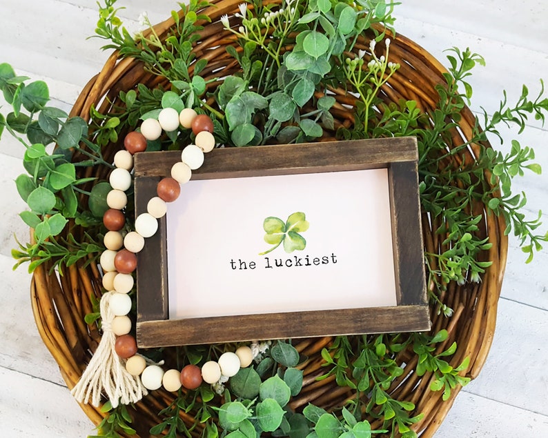 May include: A wooden sign with the text "the luckiest" and a watercolor illustration of a four-leaf clover. The sign is surrounded by a wreath of green leaves and a strand of wooden beads with a tassel.
