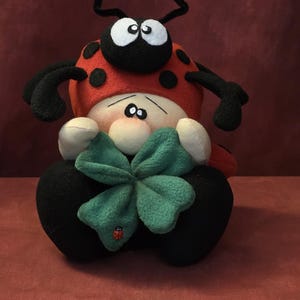 May include: A soft toy ladybird doll with a red body, black spots, and a black head with white eyes and antennae. It holds a green four-leaf clover with a small ladybird applique. The doll is sitting on a red surface.
