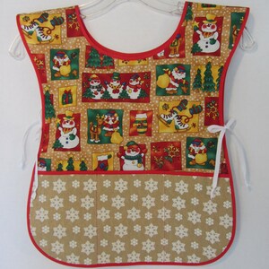 Puede incluir: Un delantal de Navidad rojo y marr&oacute;n con un dise&ntilde;o de mu&ntilde;eco de nieve y copos de nieve. El delantal tiene un bolsillo grande en la parte delantera con un patr&oacute;n de copos de nieve.