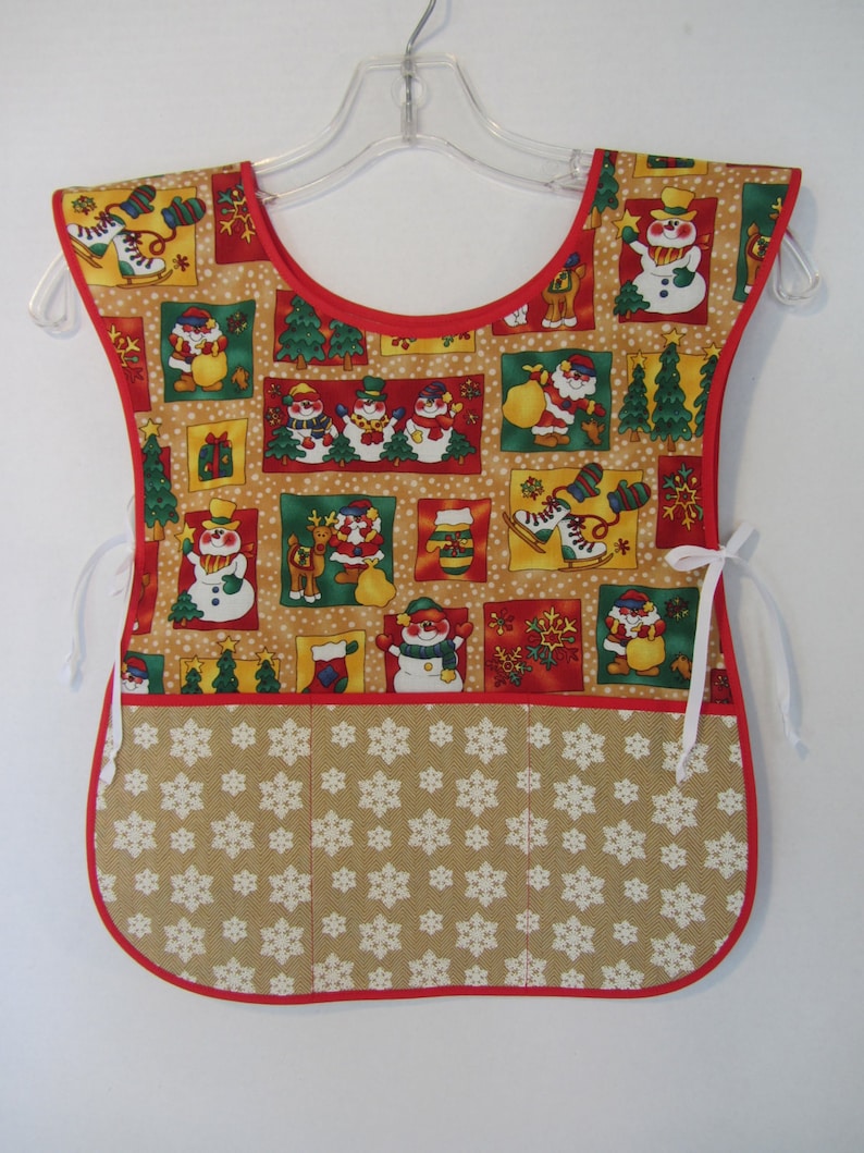 Puede incluir: Un delantal de Navidad rojo y marr&oacute;n con un dise&ntilde;o de mu&ntilde;eco de nieve y copos de nieve. El delantal tiene un bolsillo grande en la parte delantera con un patr&oacute;n de copos de nieve.