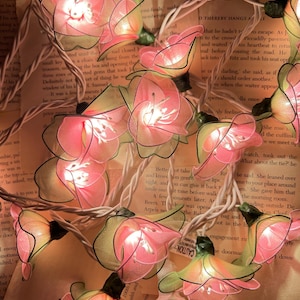May include: A string of decorative fairy lights featuring pink and yellow fabric flower blossoms. The lights are strung on a white wire and are illuminated, creating a warm glow. The flowers are arranged on a background of open book pages.