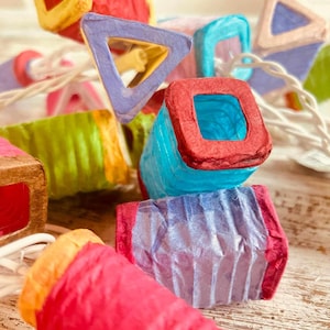 K&ouml;nnte beinhalten: Eine Lichterkette mit bunten Papierlaternen in verschiedenen Formen, darunter Quadrate, Dreiecke und Zylinder. Die Laternen sind mit wei&szlig;em Draht verbunden und strahlen ein warmes, einladendes Licht aus.