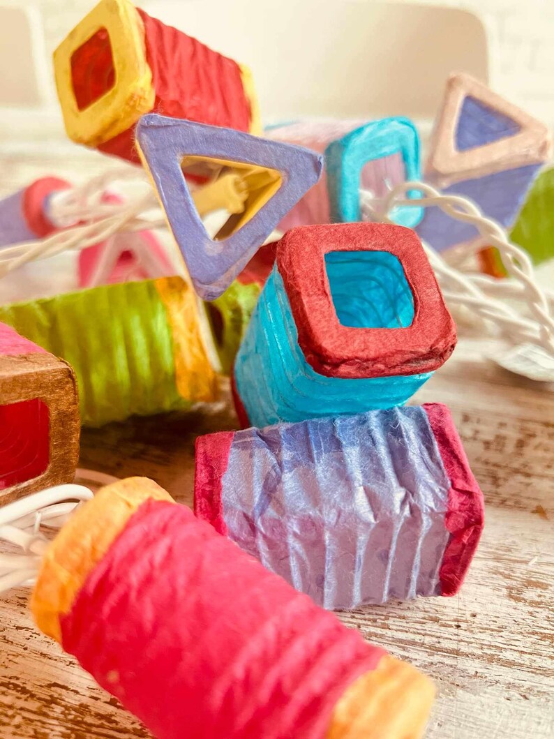 K&ouml;nnte beinhalten: Eine Lichterkette mit bunten Papierlaternen in verschiedenen Formen, darunter Quadrate, Dreiecke und Zylinder. Die Laternen sind mit wei&szlig;em Draht verbunden und strahlen ein warmes, einladendes Licht aus.
