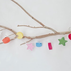 May include: A string of lights with wooden shapes, including stars, moons, and circles, in various colors, hanging from a branch.