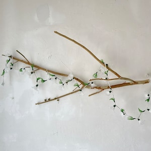 May include: White string lights with green leaves shaped lights strung on a bare branch against a white wall.