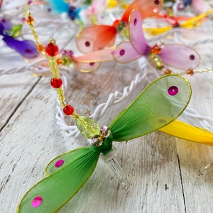 May include: A string of fairy lights with dragonfly shapes made of sheer fabric in various colors, including green, yellow, pink, orange, and blue. Each dragonfly has a small, clear light bulb and is accented with beads.