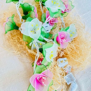 Puede incluir: Una cadena de luces de hadas con bombillas en forma de flores blancas y rosas. Las luces están envueltas alrededor de un lecho de material similar a la paja.