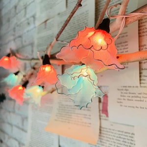 Puede incluir: Una cadena de luces decorativas con pantallas en forma de flor en tonos naranja, rosa y azul. Las luces están ensartadas en un cable fino y sujetas a una rama natural. El fondo presenta páginas de un libro y una pared de ladrillos blancos.