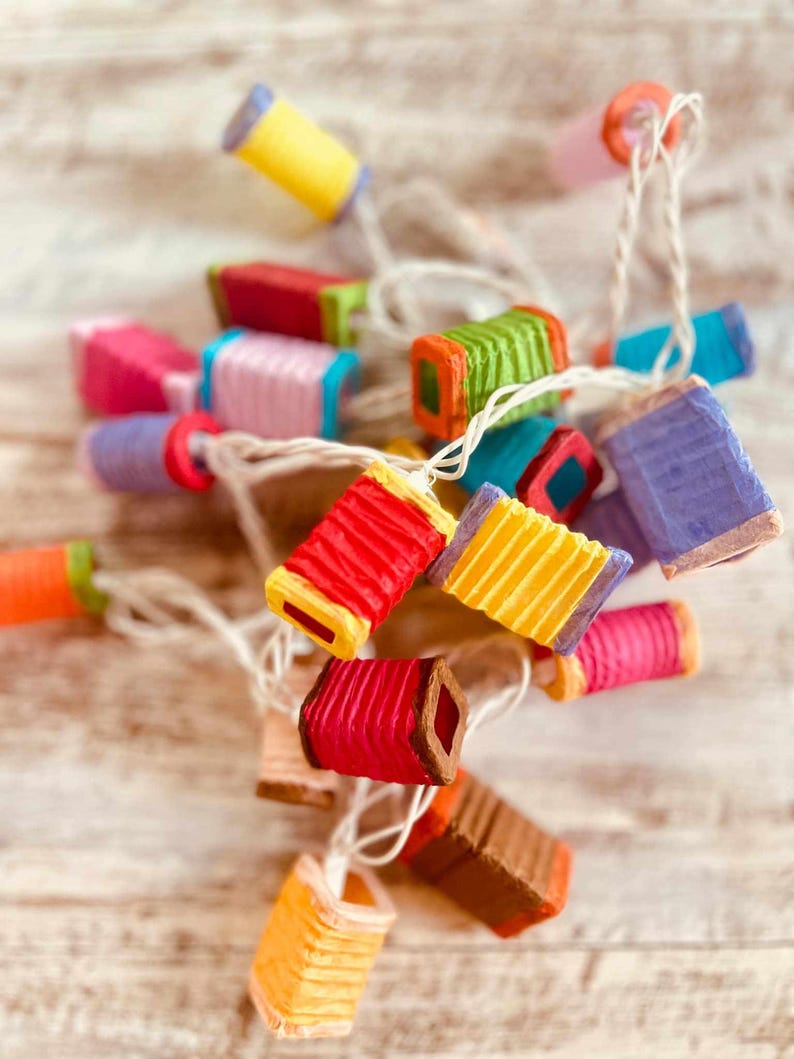 K&ouml;nnte beinhalten: Eine Lichterkette mit bunten Papierlaternen und einem wei&szlig;en Kabel. Die Laternen haben verschiedene Formen und Gr&ouml;&szlig;en, einige haben ein gefaltetes Design.