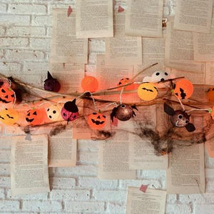 May include: A Halloween-themed string light garland with orange pumpkin and bat-shaped lights. The lights are strung on a branch, with a backdrop of vintage book pages attached to a white brick wall.