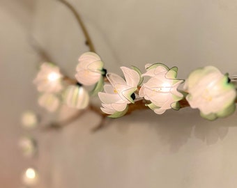 May include: A string of white paper flower lights with green accents, strung on a branch, against a white wall.