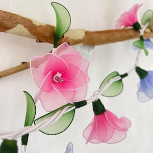 Puede incluir: Una cadena de luces con luces en forma de flores rosas y azules. Las luces están enhebradas en un cable blanco y están decoradas con hojas verdes.