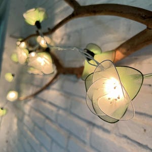 May include: Decorative string lights featuring flower-shaped shades in white and green, illuminated by small bulbs. The lights are attached to a brown branch, creating a natural and whimsical aesthetic. The background is a white brick wall.