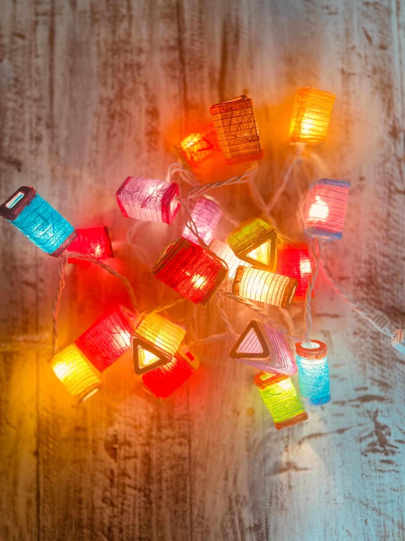 K&ouml;nnte beinhalten: Eine Lichterkette mit bunten Papierlaternen und warmwei&szlig;en Lichtern. Die Laternen haben verschiedene Formen und Gr&ouml;&szlig;en, darunter Quadrate, Dreiecke und Rechtecke. Die Lichter sind mit einem durchsichtigen Draht verbunden.