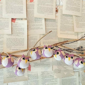 May include: A garland of small, round owl ornaments in shades of purple, white, and yellow. The owls are strung on a thin, natural-colored branch, set against a backdrop of vintage book pages.