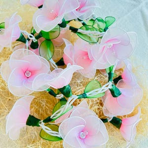 May include: A string of fairy lights with pink flower-shaped bulbs and green leaves. The lights are wrapped around a bed of straw-like material.