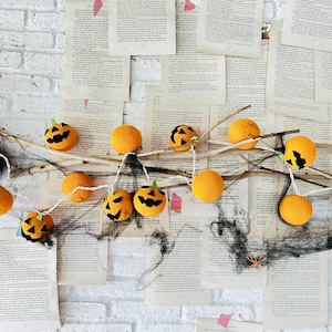 Puede incluir: Una guirnalda de calabazas de fieltro naranja con caras negras talladas y tallos verdes, ensartadas en un cordón blanco. Las calabazas están dispuestas sobre ramas sobre un fondo de páginas de libros antiguos y una pared de ladrillos blancos.