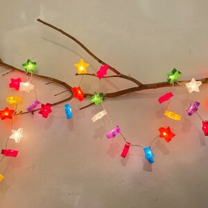May include: A string of colorful star-shaped lights, in shades of red, green, yellow, and blue, are strung along a thin wire. The lights are draped over a brown branch against a neutral wall. The stars are lit, creating a warm, festive glow.