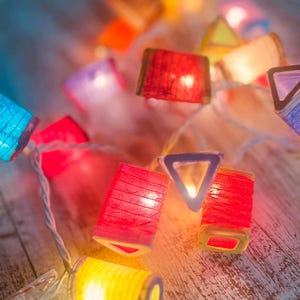 K&ouml;nnte beinhalten: Eine Lichterkette mit bunten Papierlaternen, in denen Lichter brennen. Die Laternen haben verschiedene Formen, darunter Quadrate, Dreiecke und Rechtecke. Die Lichter leuchten und verbreiten ein warmes, einladendes Licht.