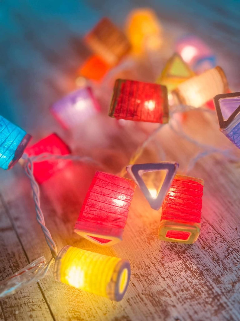 K&ouml;nnte beinhalten: Eine Lichterkette mit bunten Papierlaternen, in denen Lichter brennen. Die Laternen haben verschiedene Formen, darunter Quadrate, Dreiecke und Rechtecke. Die Lichter leuchten und verbreiten ein warmes, einladendes Licht.