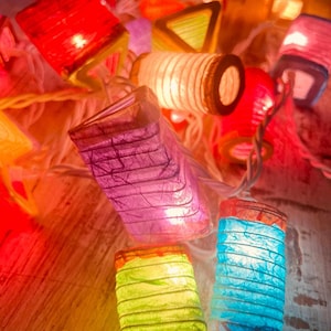 K&ouml;nnte beinhalten: Eine Lichterkette mit bunten Papierlaternen und einem wei&szlig;en Kabel. Die Laternen haben verschiedene Formen, darunter Quadrate, Dreiecke und Zylinder. Die Laternen sind beleuchtet und leuchten.
