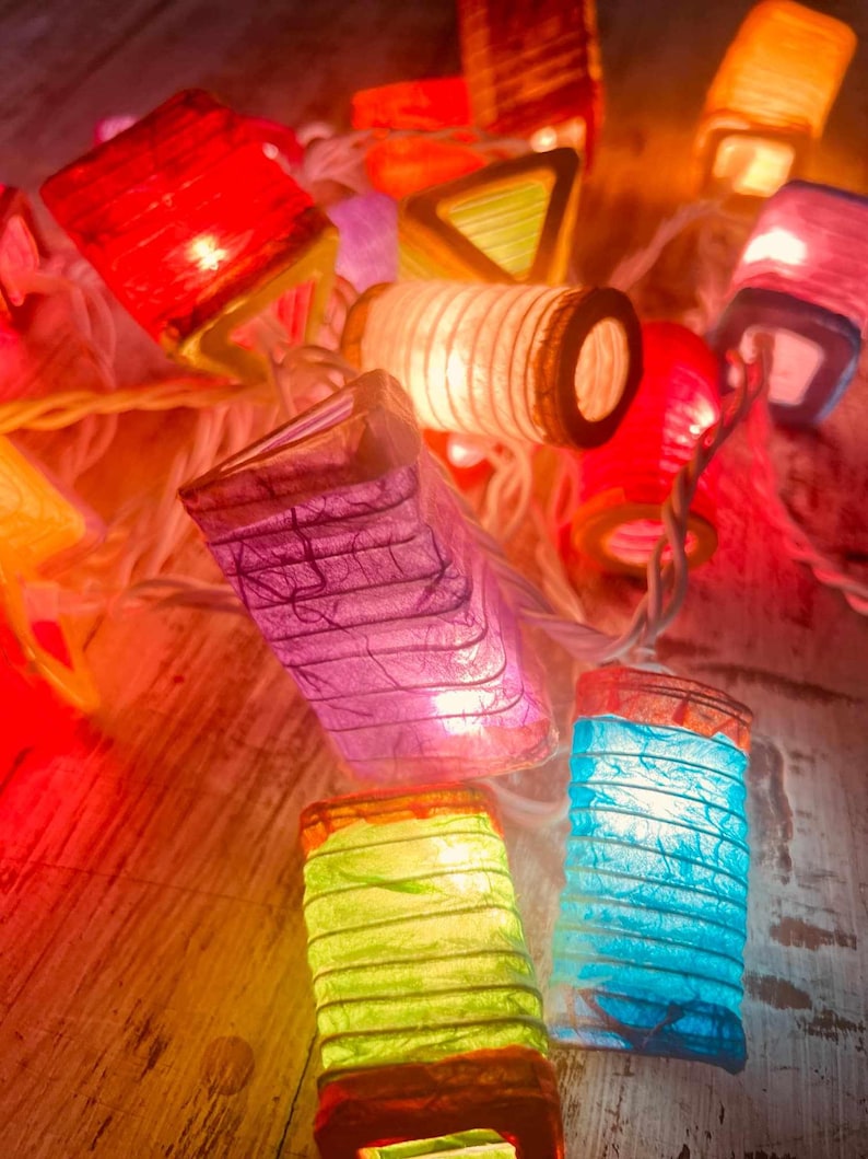 K&ouml;nnte beinhalten: Eine Lichterkette mit bunten Papierlaternen und einem wei&szlig;en Kabel. Die Laternen haben verschiedene Formen, darunter Quadrate, Dreiecke und Zylinder. Die Laternen sind beleuchtet und leuchten.