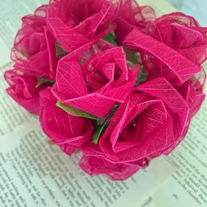 May include: A bouquet of vibrant pink artificial roses. The roses are clustered together, with delicate, veined petals. The bouquet rests on the pages of an open book, creating a contrast of textures and colors. The overall theme is floral and decorative.